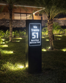 Placa residencial Malta preta com LED aceso à noite em jardim com palmeiras

